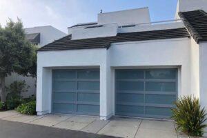 A modern white house with two gray garage doors and green shrubs on a clear day.